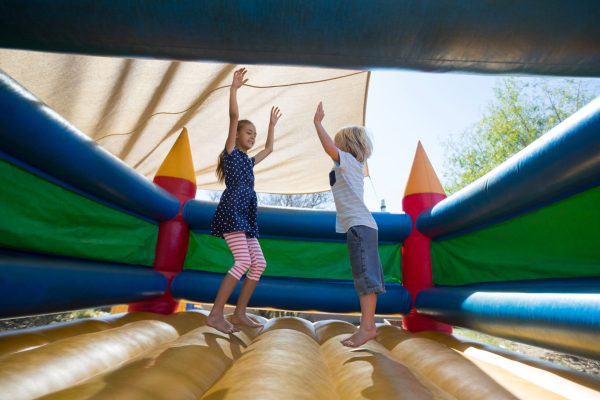 Cheerful siblings with arms raised jumping on bouncy castle at playground